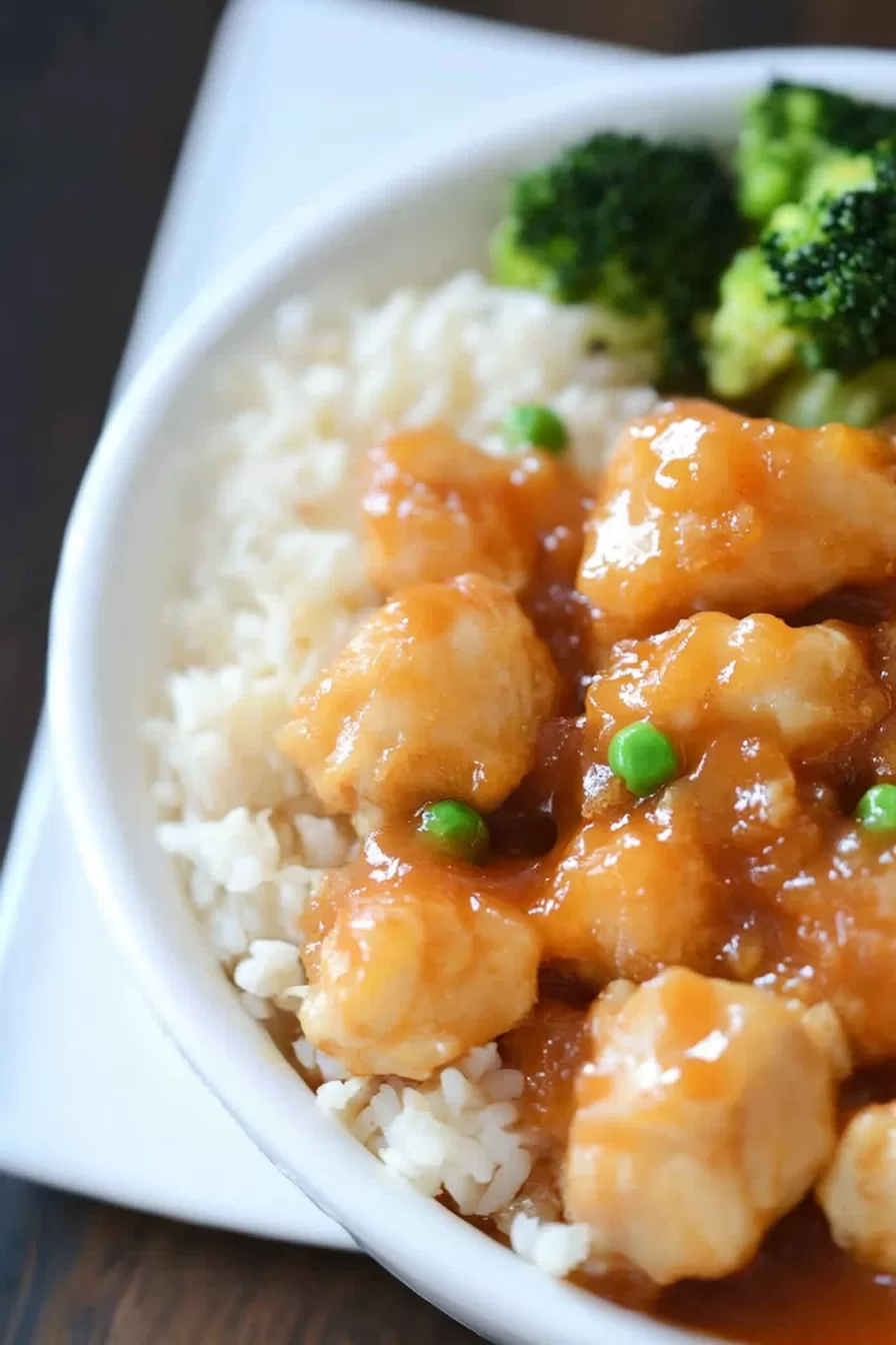 Bowl of tender sweet and sour chicken garnished with peas, paired with rice.