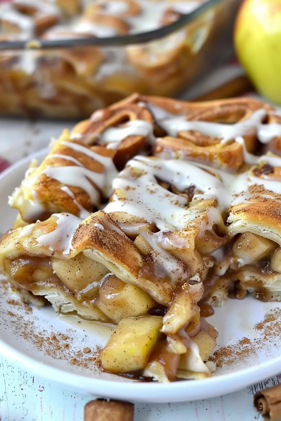 A slice of layered caramel apple cinnamon roll lasagna served on a white plate, topped with creamy icing.