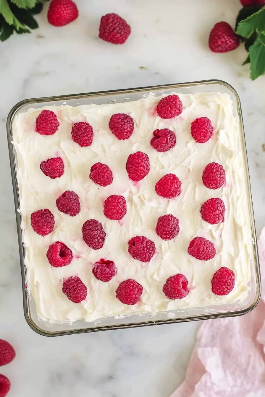 Overhead view of a coconut poke cake in a pan, decorated with evenly spaced fresh raspberries.