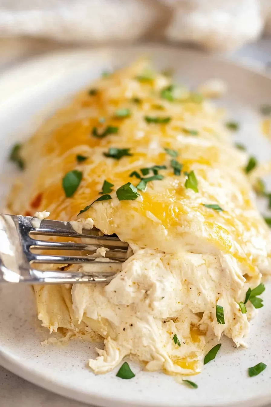 A fork revealing tender, shredded chicken filling inside a cheesy sour cream enchilada on a white plate.