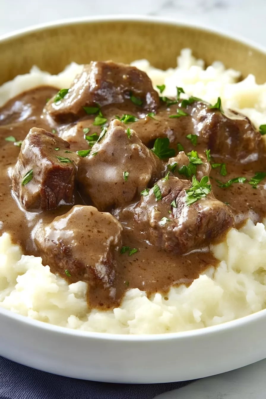 Thick, juicy beef chunks drenched in savory brown gravy, plated with mashed potatoes in a deep bowl.