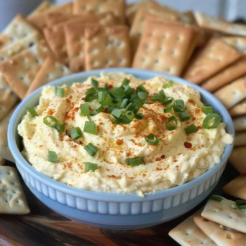 Smooth and fluffy deviled eggs dip garnished with paprika and chives, surrounded by golden crackers for dipping.