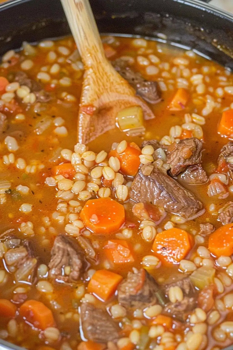 A pot of freshly cooked beef barley soup with a wooden spoon stirring through beef pieces, carrots, and barley grains in a savory broth.