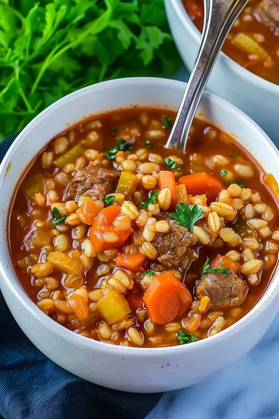 A spoon scooping a hearty serving of beef barley soup from a bowl, highlighting the beef chunks, barley grains, and vegetables in a rich tomato-based broth.