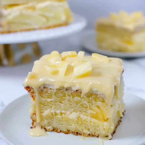 A slice of pineapple cake dripping with creamy frosting and topped with fresh pineapple chunks on a white plate.