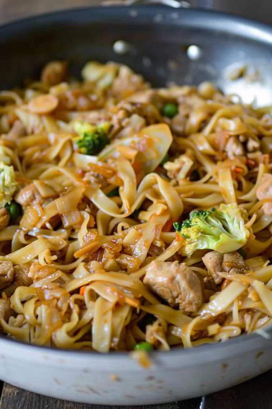 Close-up of a skillet filled with tender noodles, vegetables, and juicy chicken coated in a savory sauce.