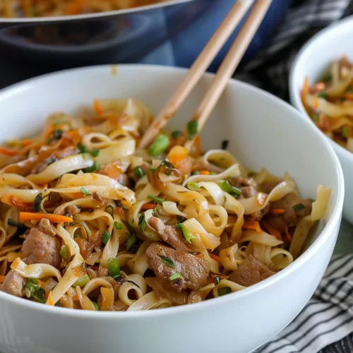 Chopsticks lifting a portion of noodle stir-fry, showing the saucy noodles, chicken pieces, and crisp vegetables.