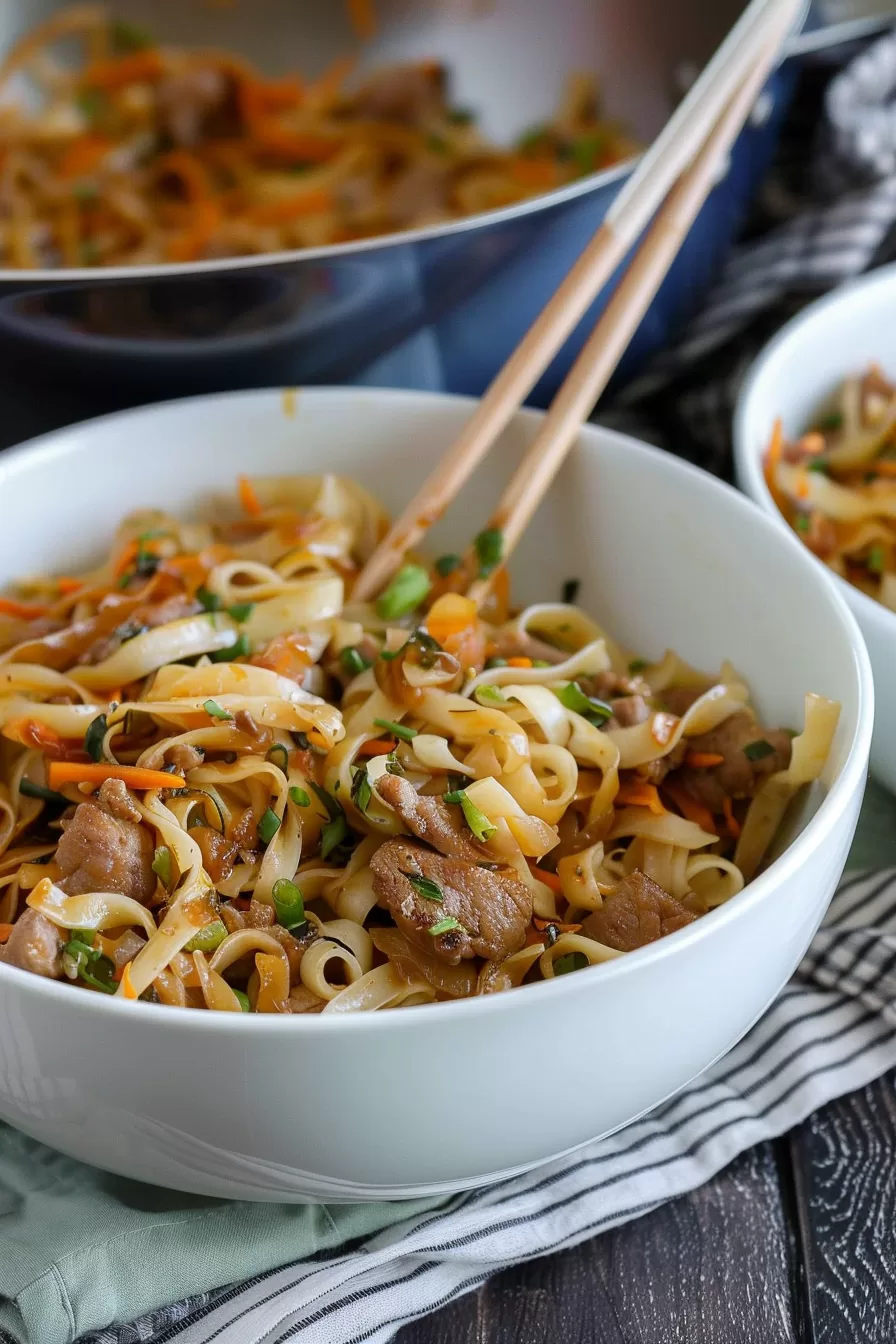 Chopsticks lifting a portion of noodle stir-fry, showing the saucy noodles, chicken pieces, and crisp vegetables.