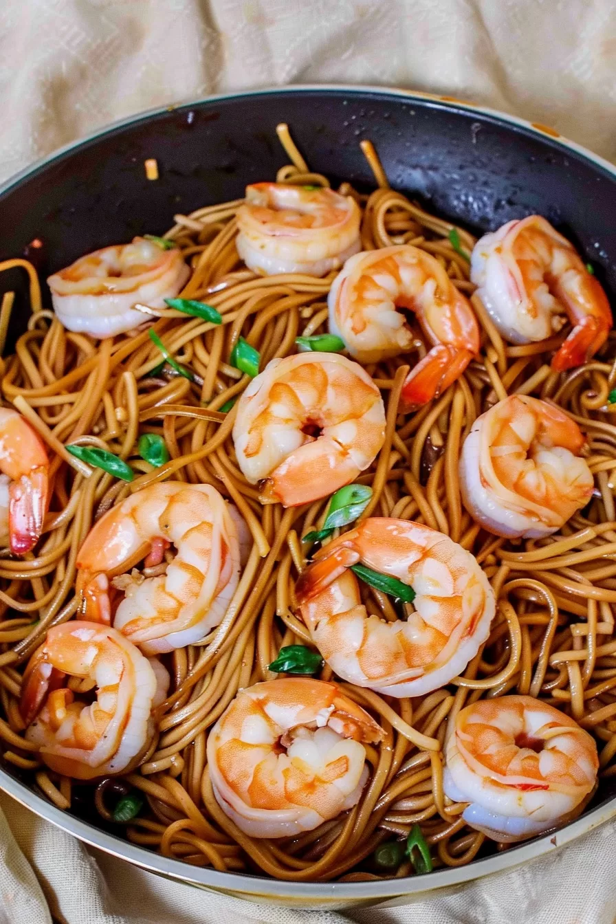 Top-down view of shrimp lo mein in a skillet, highlighting the plump shrimp, golden noodles, and fresh garnishes with a rich sauce.