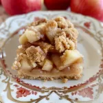 A serving of homemade apple pie bars on a decorative plate with fresh apples.