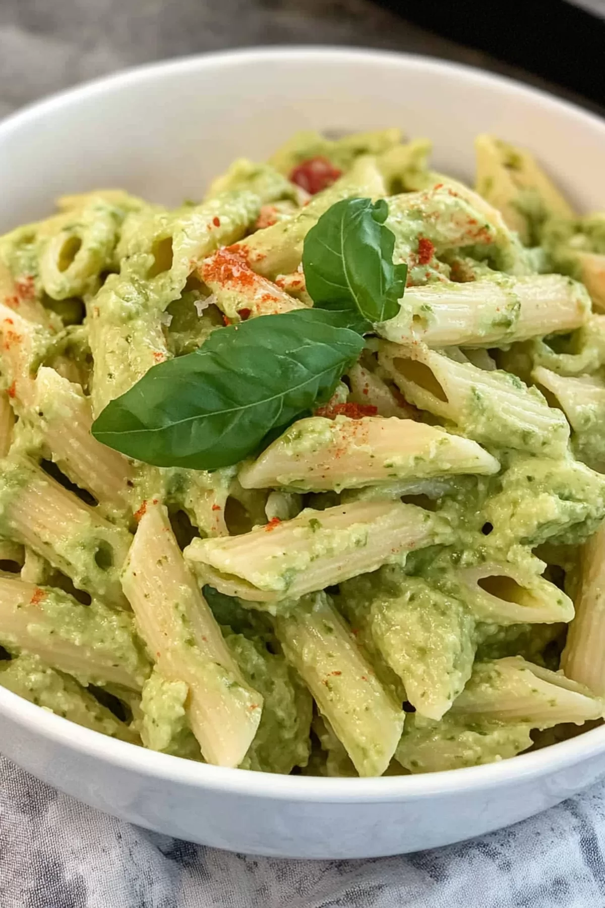 A bowl of pasta tossed in a vibrant green, creamy avocado sauce, garnished with fresh herbs.