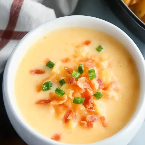 Close-up of a hearty, cheesy soup with vibrant toppings served in a clean white bowl on a wooden surface.