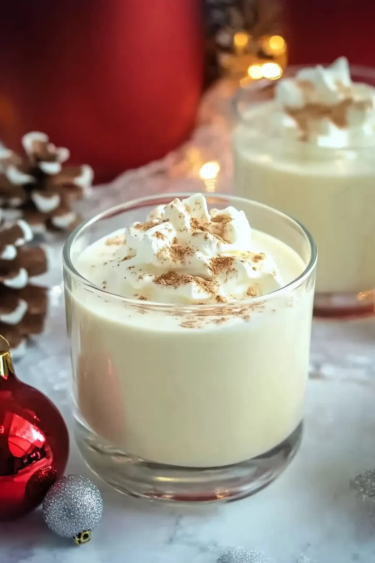 A festive glass of creamy eggnog topped with whipped cream and a sprinkle of nutmeg, surrounded by Christmas decorations.