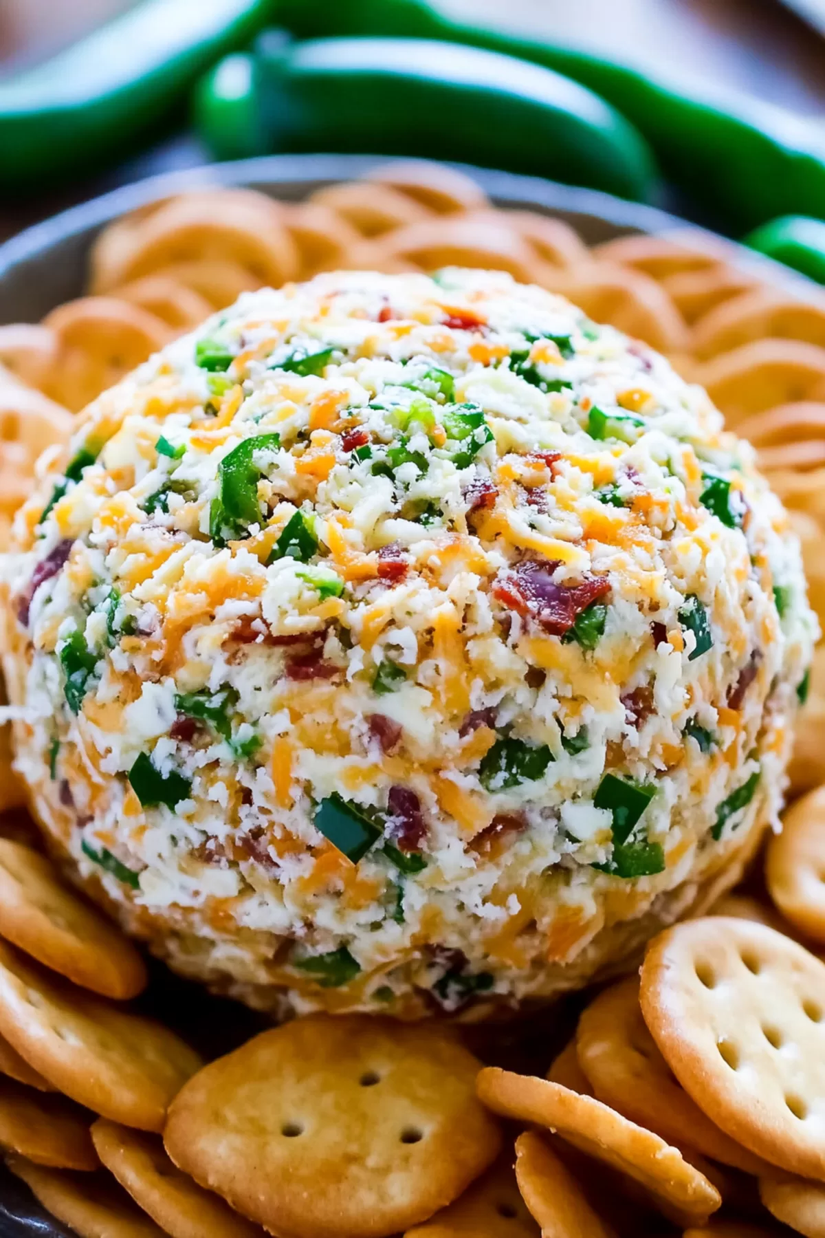 Close-up of a creamy cheese ball speckled with jalapeño, crispy bacon, and cheddar, surrounded by golden crackers on a serving dish.