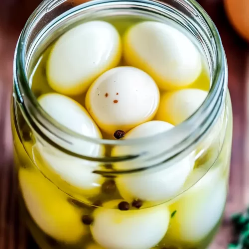 Close-up of eggs pickling in a clear glass jar, surrounded by fresh herbs and spices on a wooden countertop.