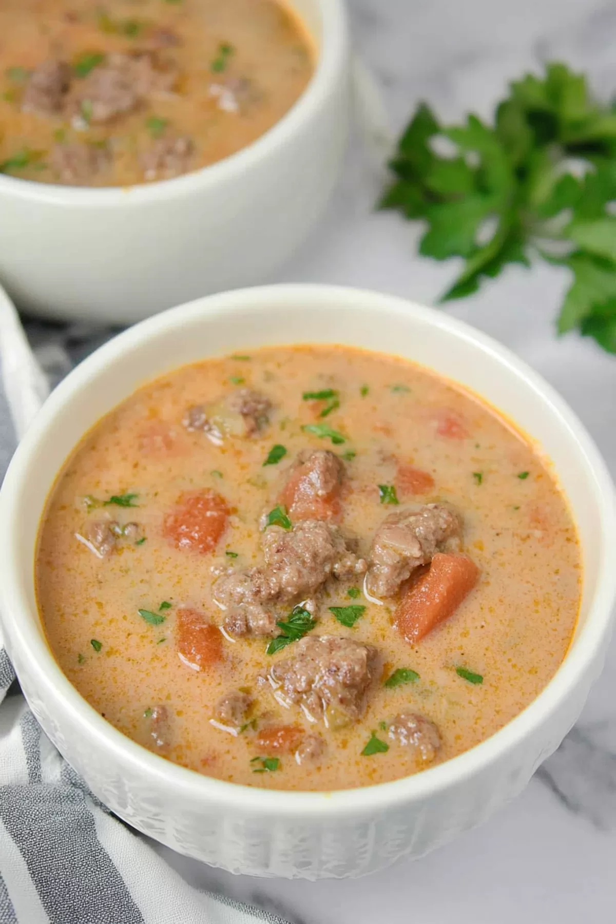 A creamy soup served in a white bowl, garnished with fresh parsley, showcasing chunks of ground beef and vibrant vegetables.