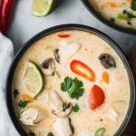 A bowl of creamy soup with a hint of spice, garnished with fresh herbs and lime slices, served on a wooden table.