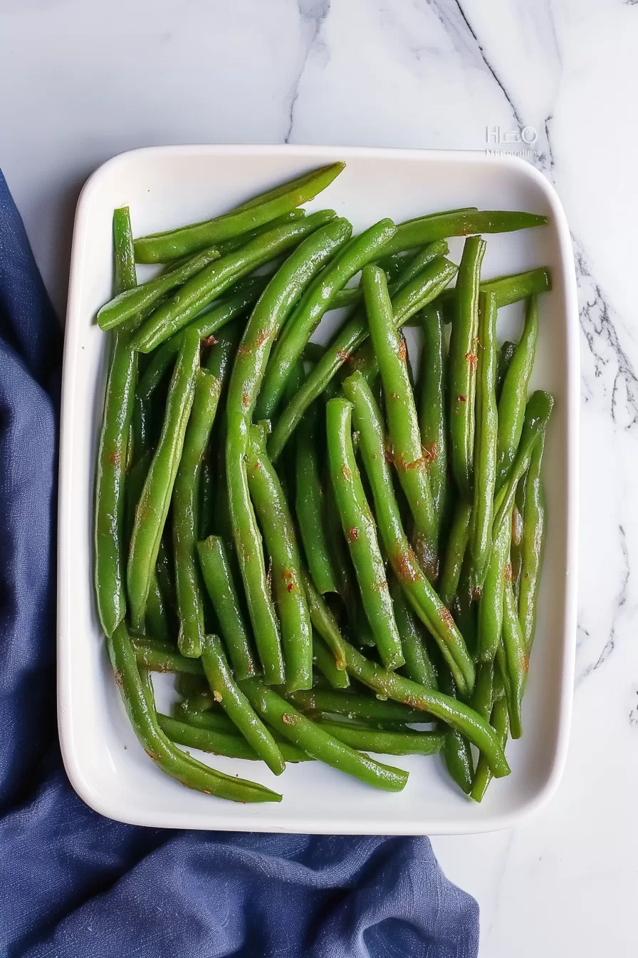 Freshly cooked green beans arranged in a white dish, glistening with a light coating of oil and seasoning.