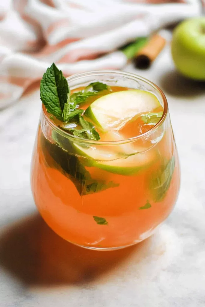 A close-up of a refreshing orange-hued drink garnished with fresh mint leaves and thin apple slices in a clear glass.