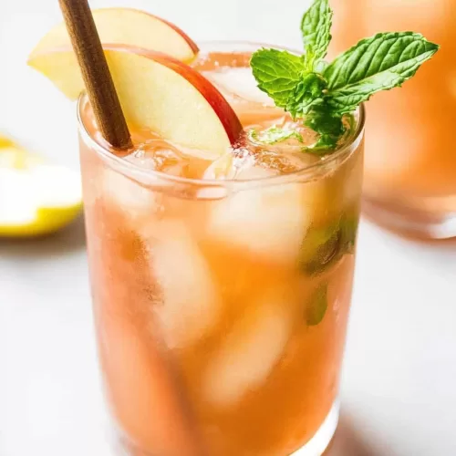 A chilled fall-inspired cocktail with visible ice cubes, fresh mint, and apple slices sitting on a white marble surface.