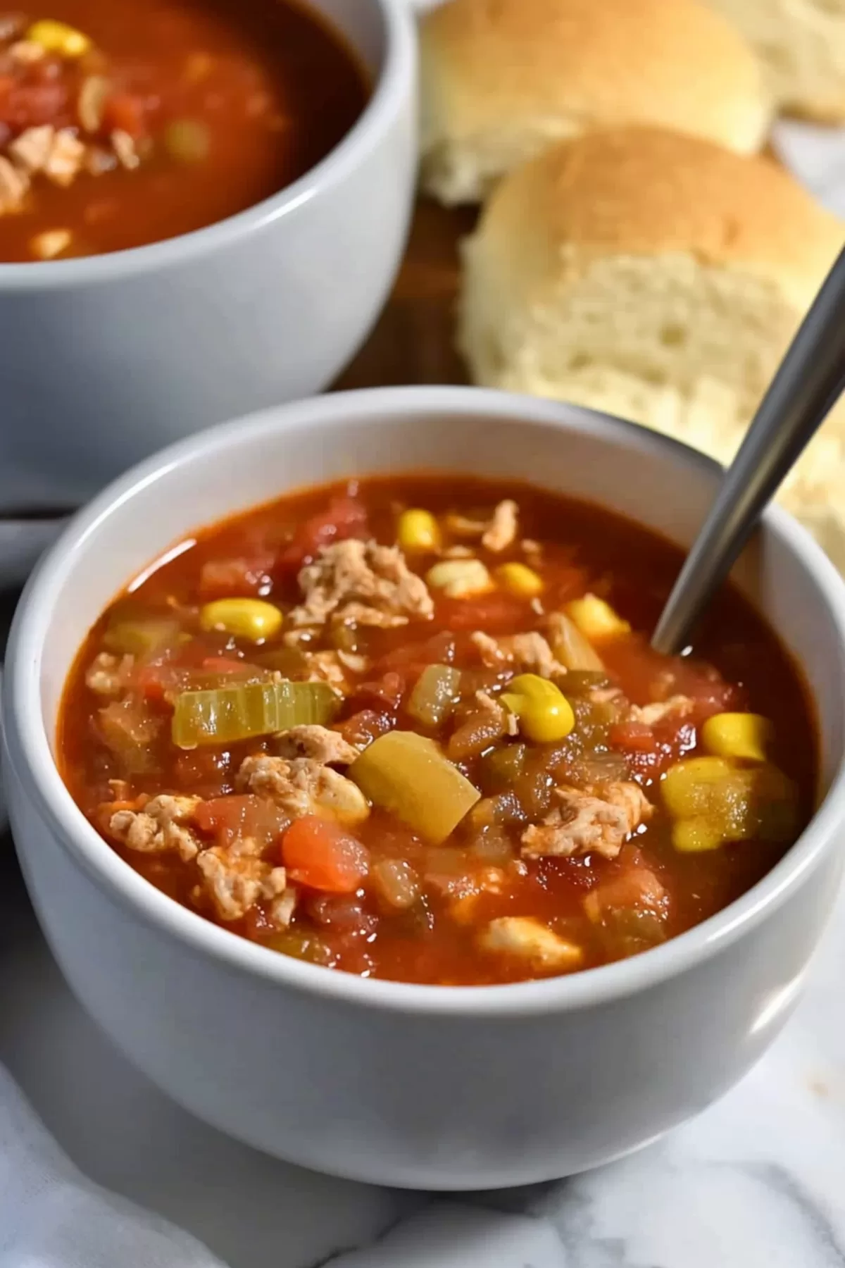 A hearty bowl of stew with a rich tomato base, corn, and tender chunks of meat served with warm bread rolls in the background.