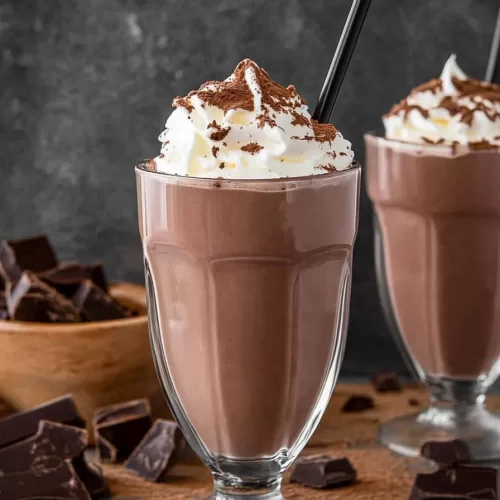 Two chocolate milkshakes with whipped cream and cocoa powder, surrounded by chocolate pieces.