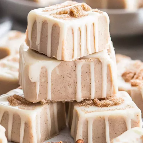 Close-up of creamy fudge squares drizzled with white icing and sprinkled with cinnamon sugar on a white plate.