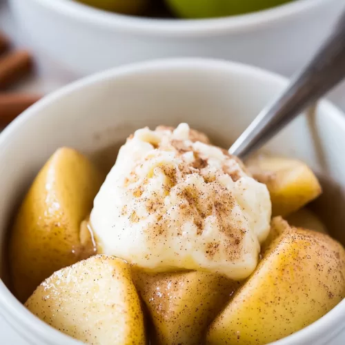 Close-up of tender cinnamon-dusted apple chunks served in a white bowl, with fresh green apples and cinnamon sticks in the background.