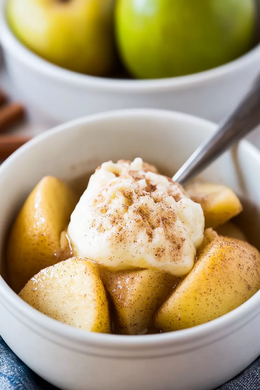 Close-up of tender cinnamon-dusted apple chunks served in a white bowl, with fresh green apples and cinnamon sticks in the background.