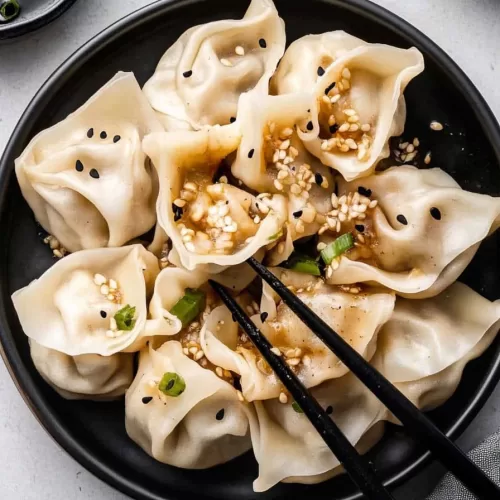 A black bowl filled with pork wontons, paired with a savory soy sauce for dipping.