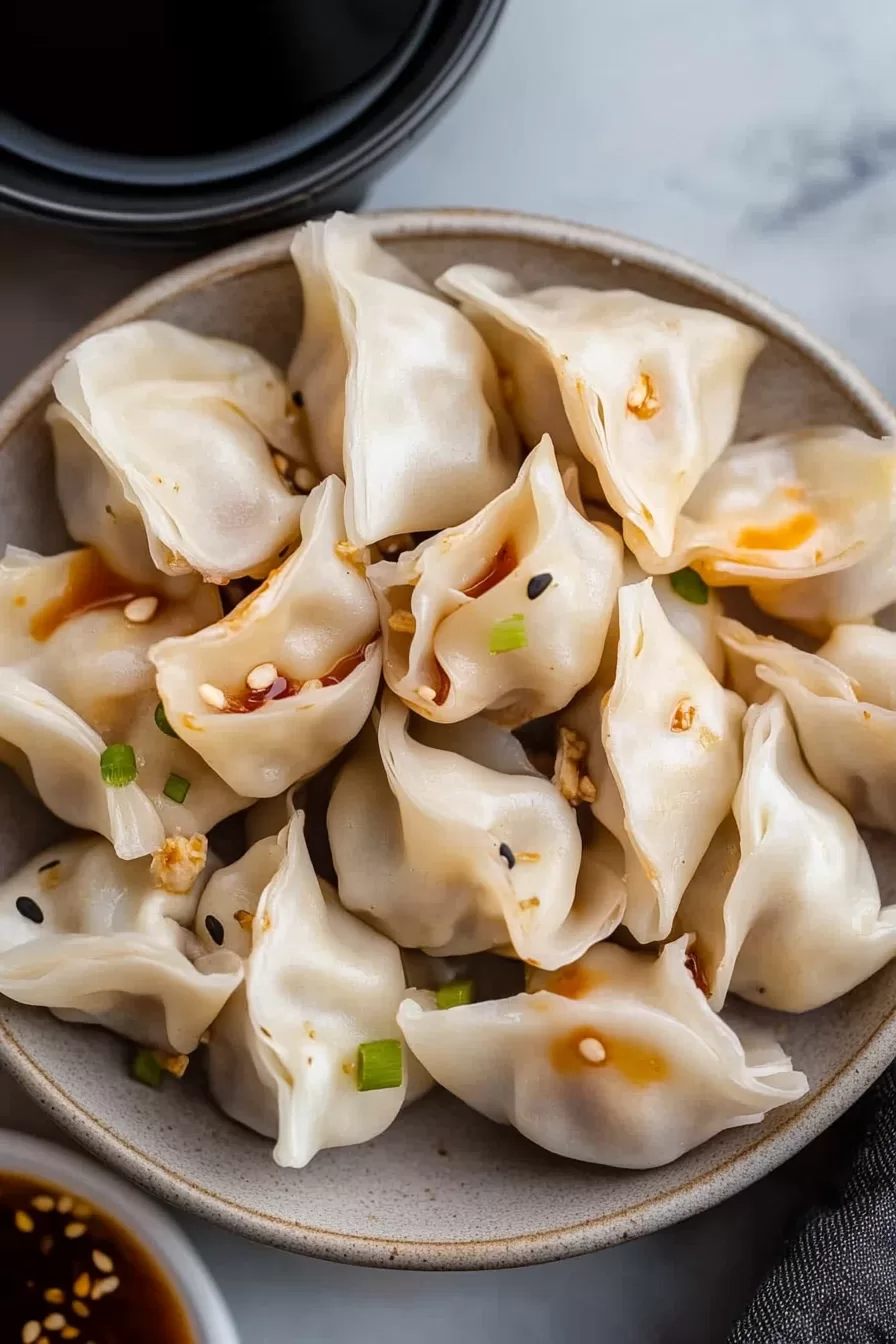 A bowl of pork wontons with a drizzle of soy-based sauce and sprinkled with sesame seeds.