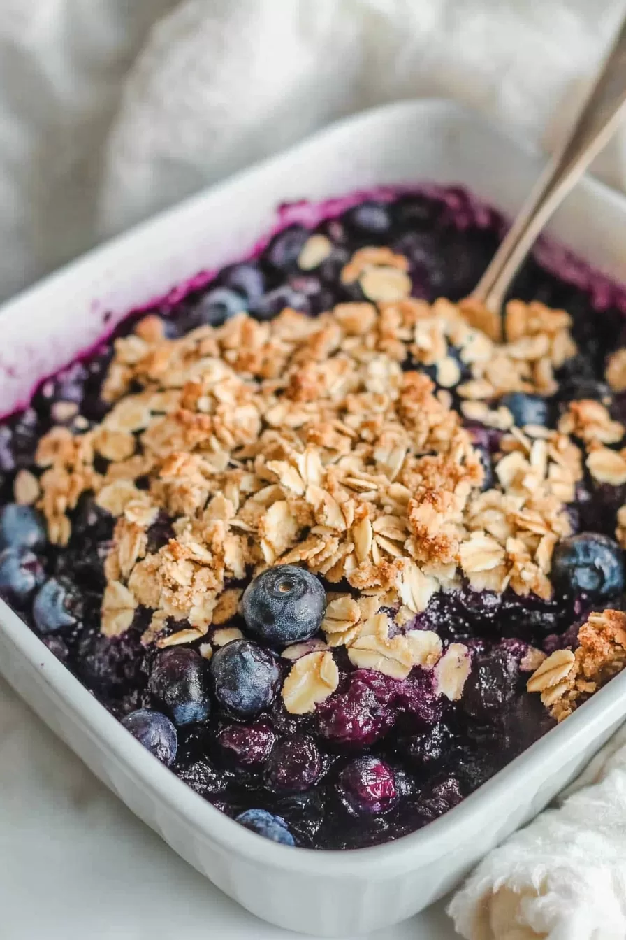 A bowl of warm blueberry crisp topped with crunchy oats and surrounded by fresh blueberries on a rustic table setting.