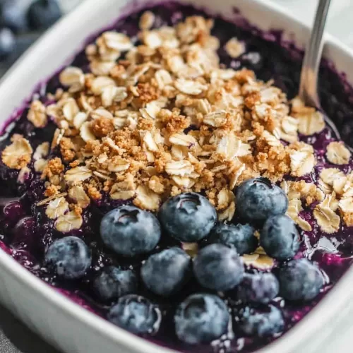 A spoon digging into a blueberry crisp, showcasing the rich, purple blueberry filling and golden crisp topping.