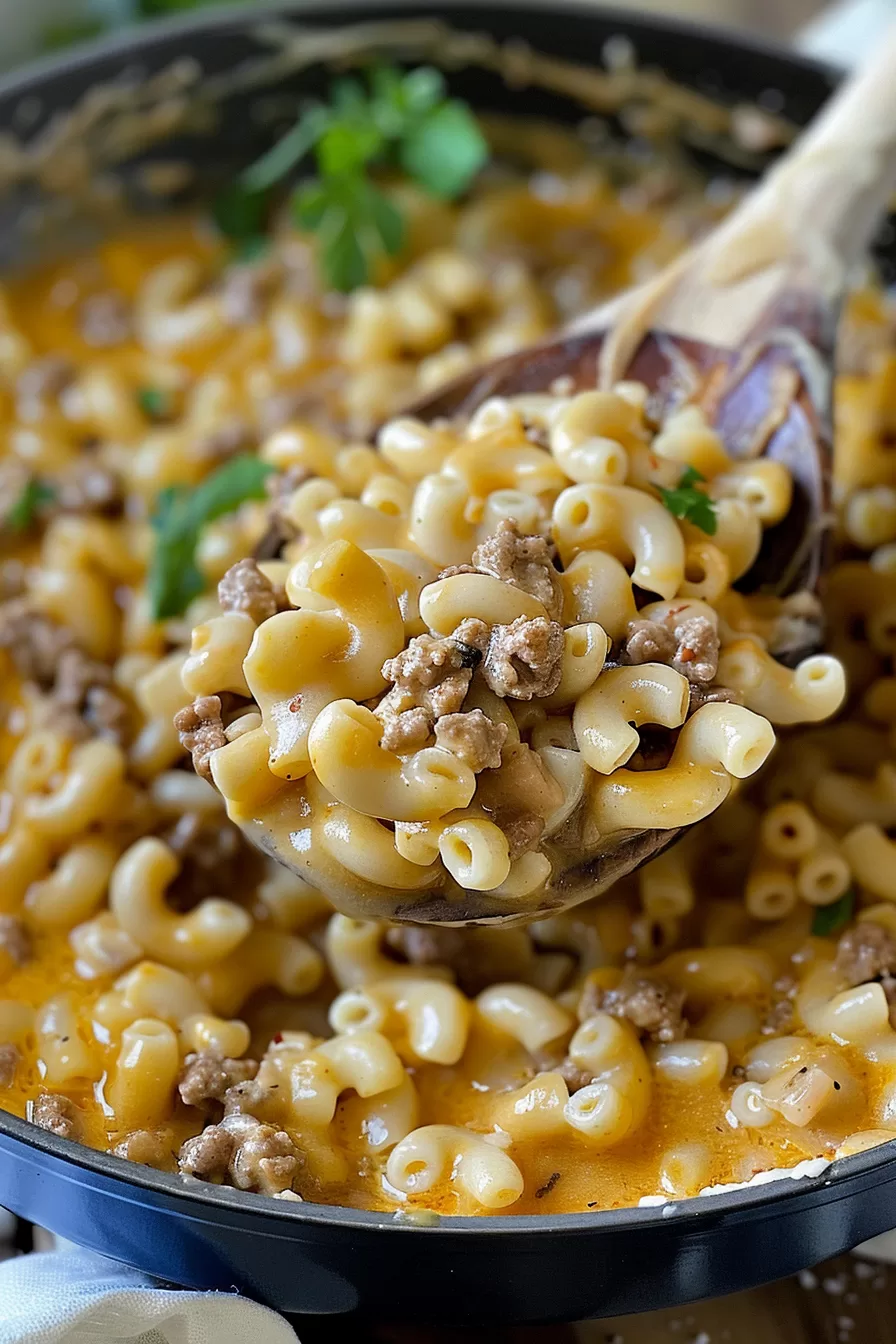 A wooden spoon scooping a portion of cheesy pasta and beef from a skillet filled with a creamy homemade hamburger helper dish.