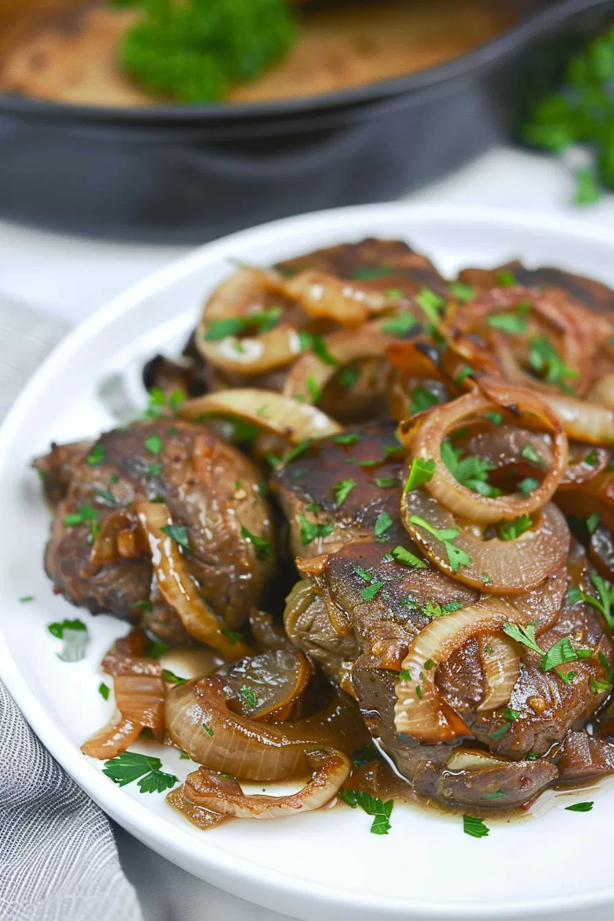 Sliced liver topped with golden-brown onions and fresh parsley, with a cast iron skillet in the background.