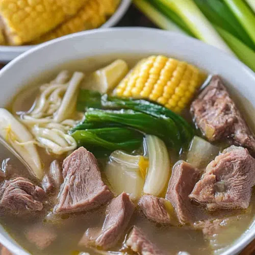 Overhead view of a hearty soup with beef shanks, corn on the cob, and leafy greens, served in a white bowl.