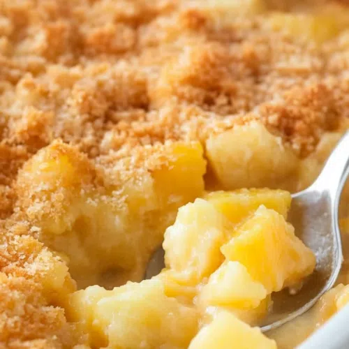 A serving spoon lifting a portion of warm, cheesy pineapple casserole from the dish.