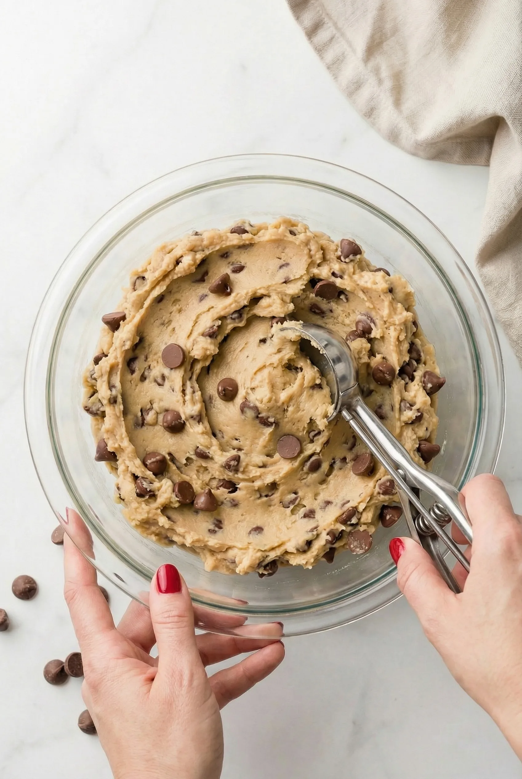 Hands holding a glass bowl while scooping chocolate chip cookie dough with a metal cookie scoop for an Edible Cookie Dough Recipe