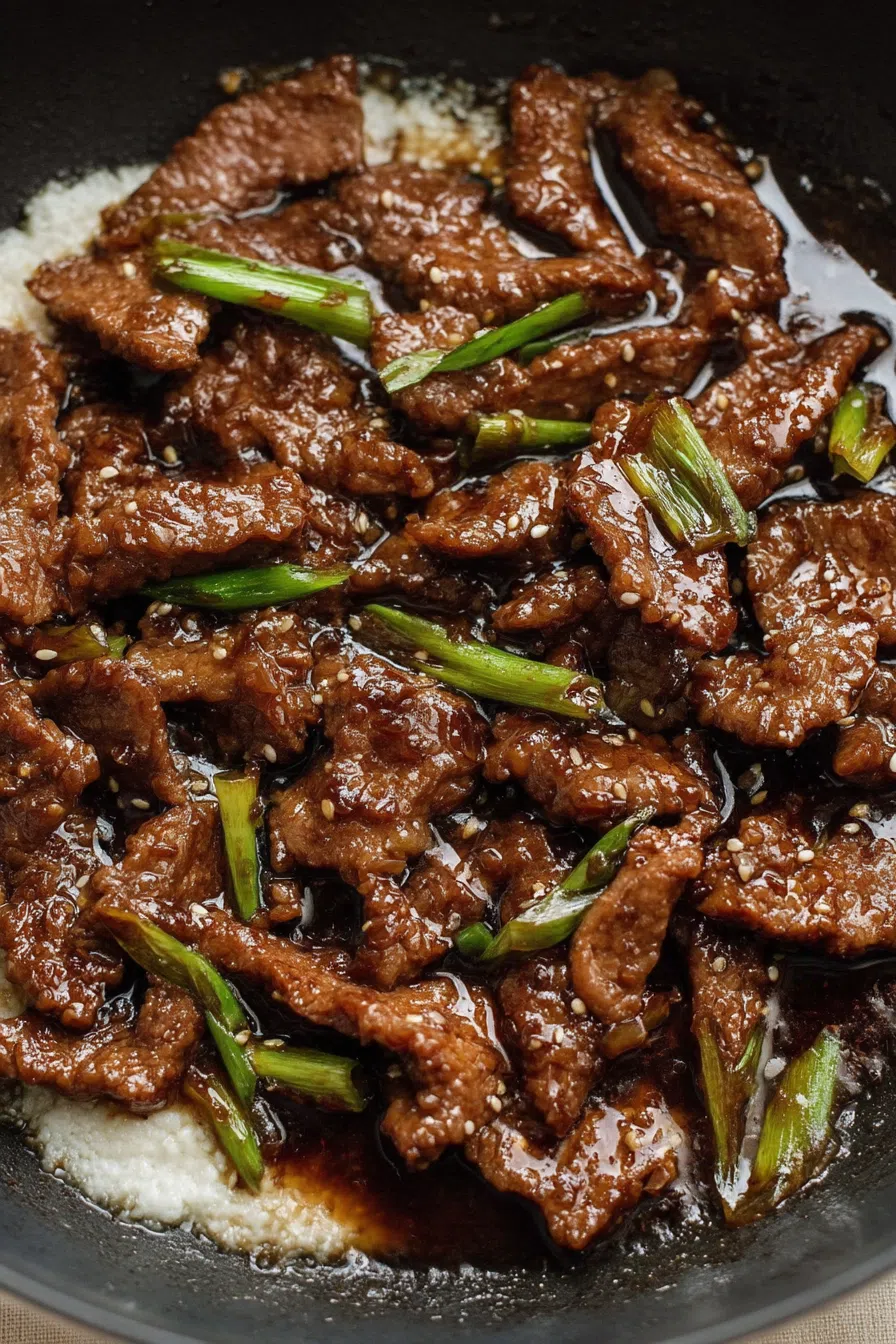 Close-up of glossy stir-fried beef strips glazed in a dark, savory sauce, garnished with chopped green onions.