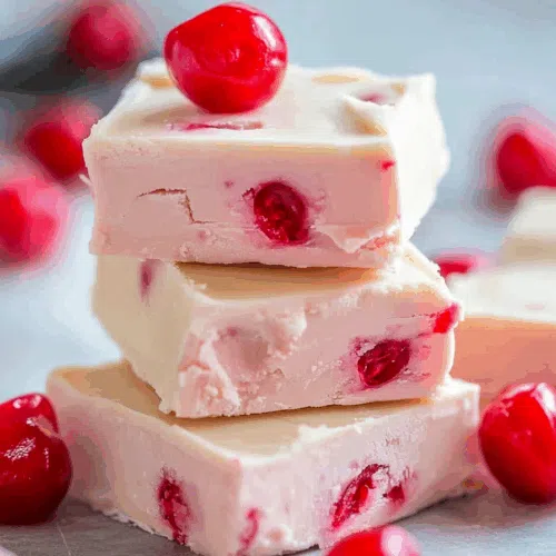 Close-up of creamy square fudge pieces with pink swirls and bits of cherry on a marble surface.