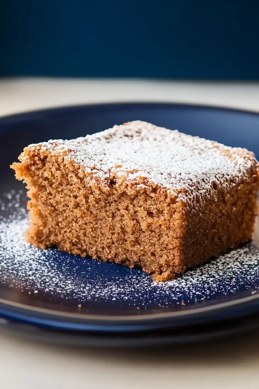 Square slice of soft, golden-brown cake dusted with powdered sugar on a navy plate.