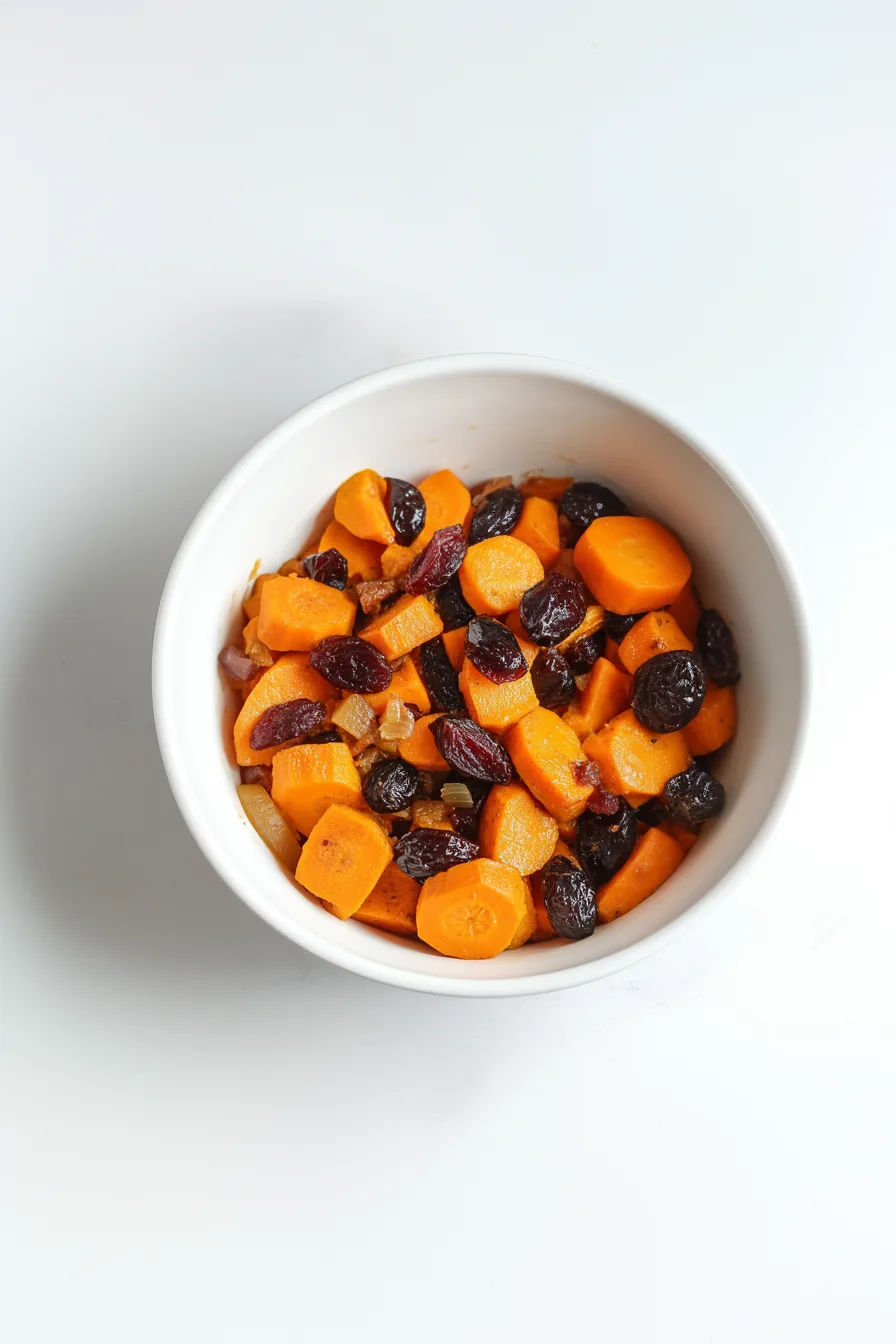 Bowl filled with glazed sweet potatoes and carrots topped with plump raisins and parsley.