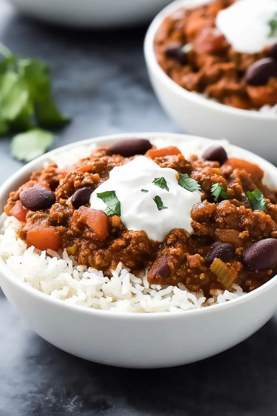 A bowl of white rice topped with a hearty bean and meat chili, finished with a dollop of sour cream.