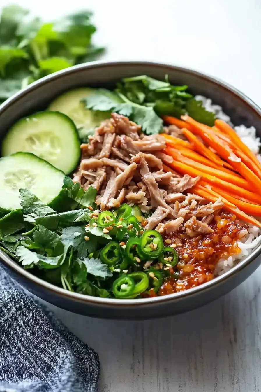 A bowl of white rice topped with tender chopped pork, sliced cucumbers, carrots, and fresh herbs.