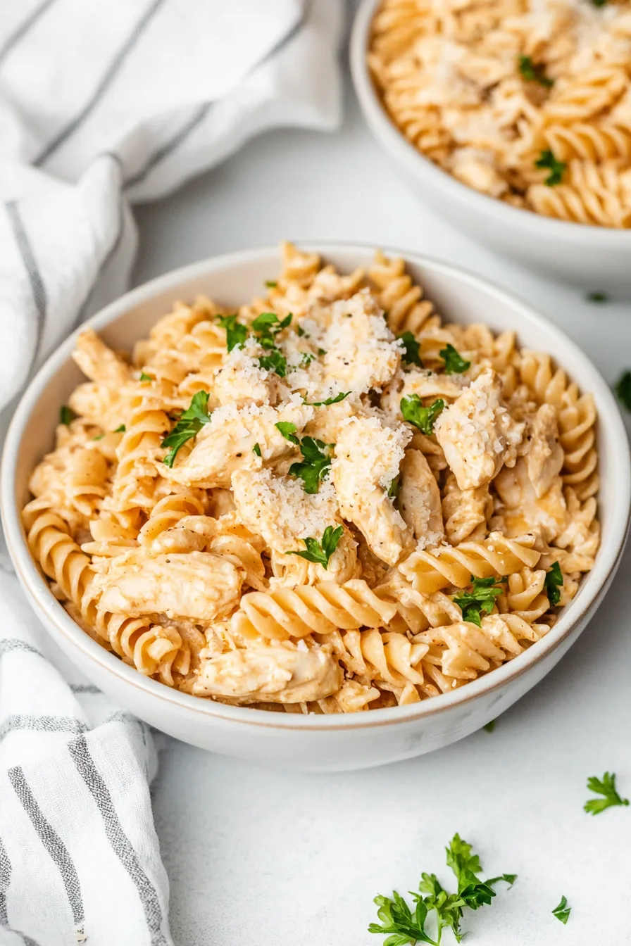 Bowl of creamy rotini pasta mixed with tender chicken and topped with grated cheese and fresh parsley.