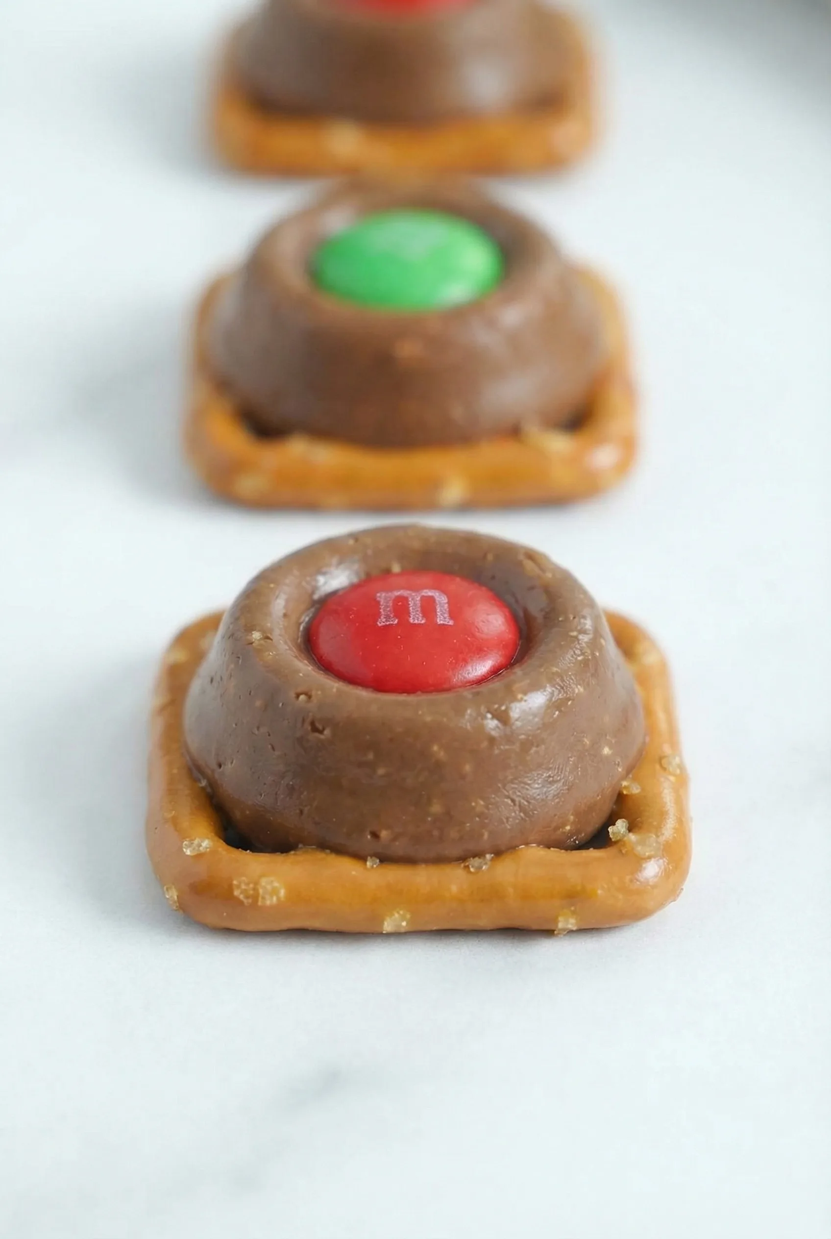 A small bite-size treat showing a soft caramel chocolate pressed onto a mini pretzel with a colorful candy on top.