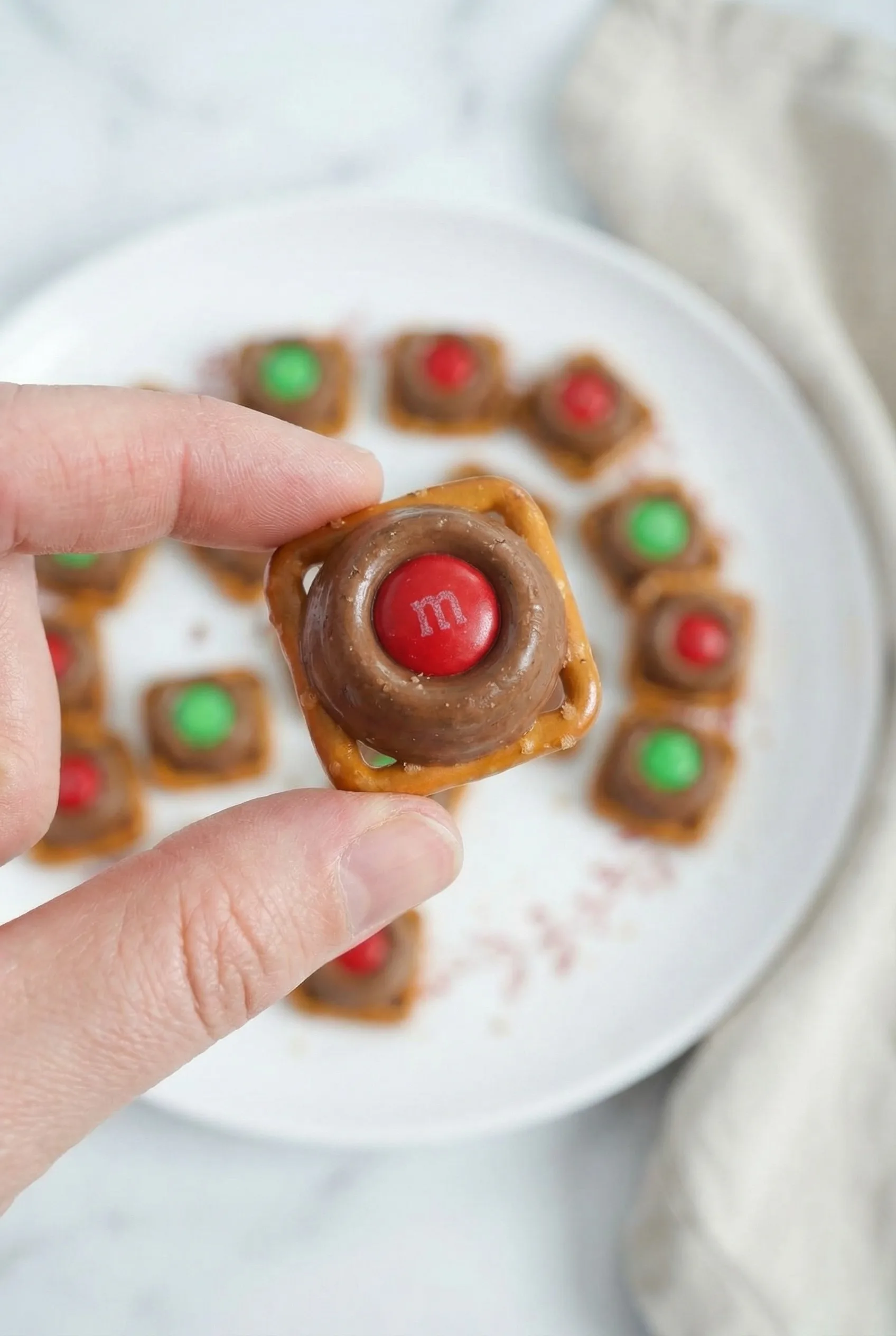 Fingers picking up a glossy caramel–chocolate pretzel bite from a plate of colorful treats.