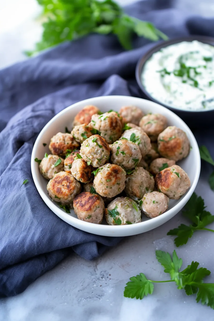 A bowl filled with seared, juicy meatballs beside a dish of herbed yogurt dip.