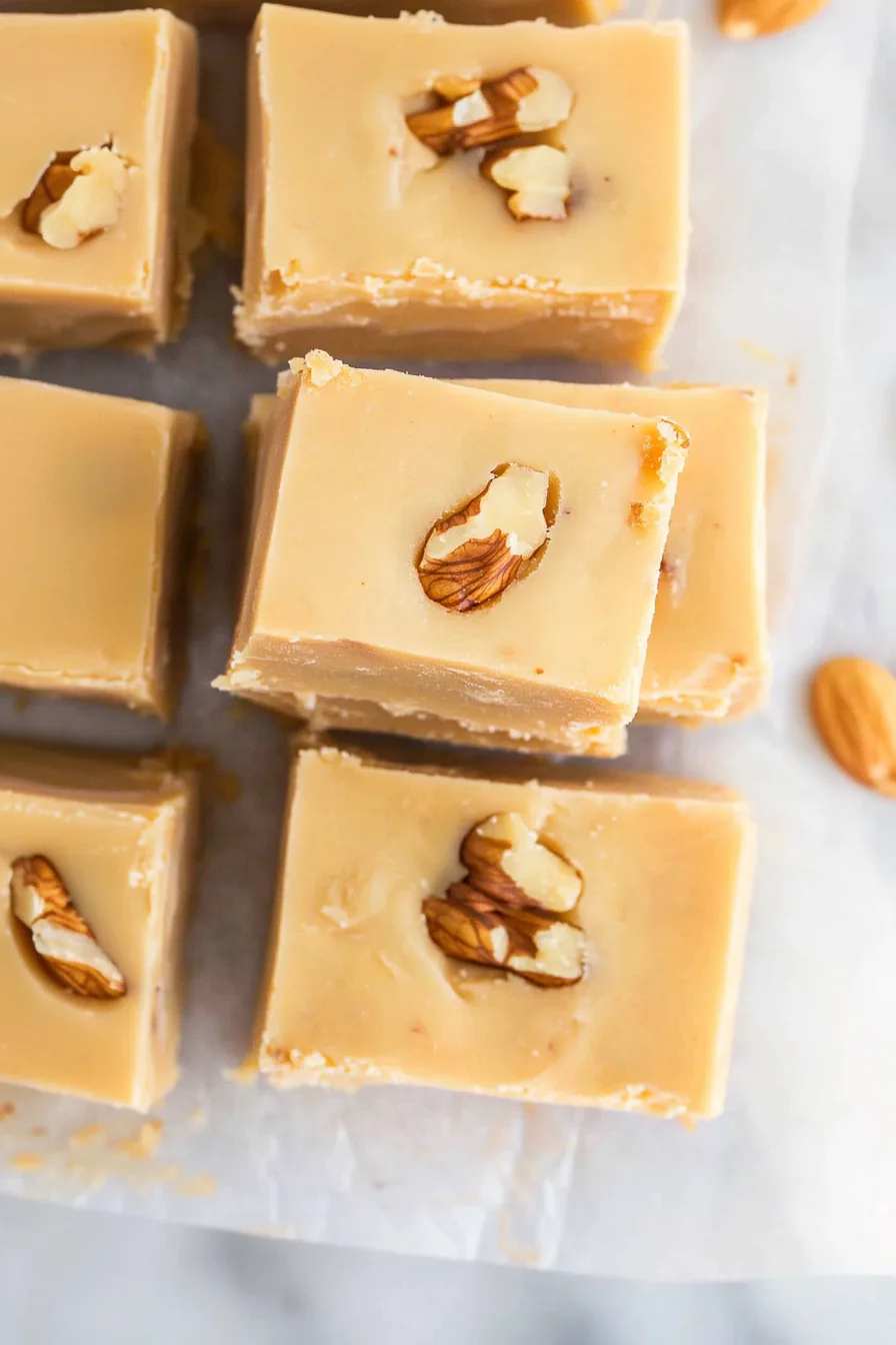 A batch of light brown fudge cubes arranged on a marble surface, each piece showing crunchy nut pieces inside.